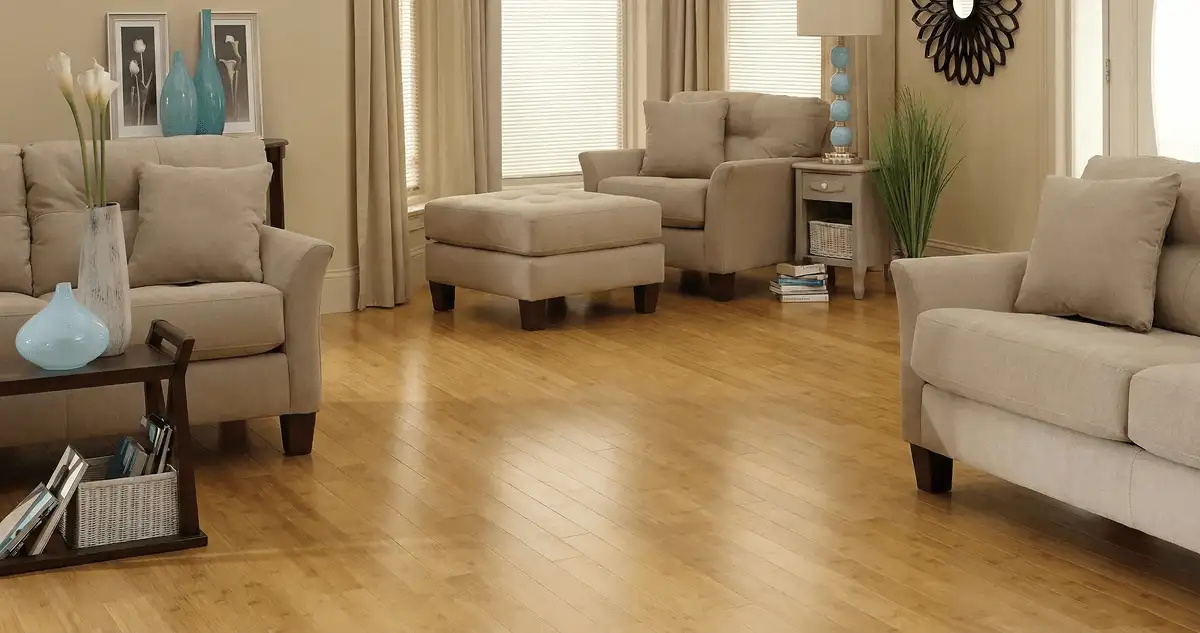 A living room with professionally installed bamboo flooring, beige sofas, a matching ottoman, a side table with a blue lamp, vases, framed pictures, and large windows with blinds and curtains in Hialeah, FL
