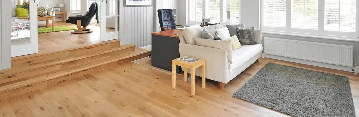 A modern living room with professionally installed laminate flooring, a beige sofa, a small side table, a gray rug, and large windows with white shutters in Hialeah, FL